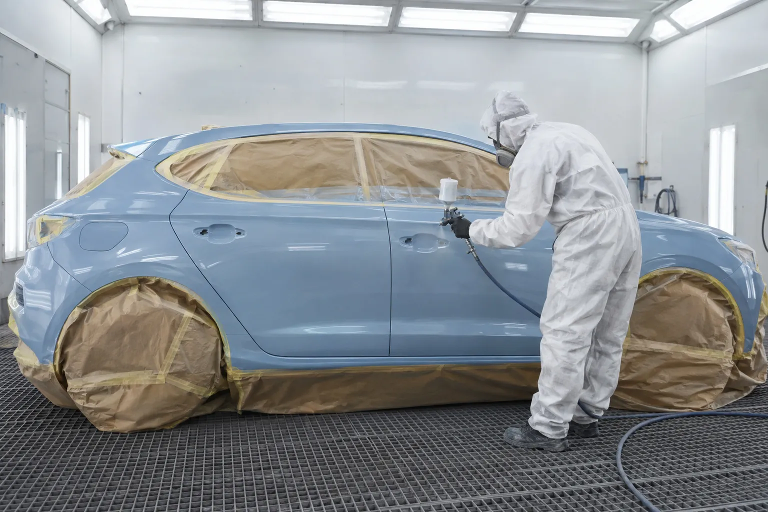 Une voiture entière dans la cabine de peinture, vitres et roues masquées, un peintre en combinaison blanche applique le vernis sur la portière arrière.